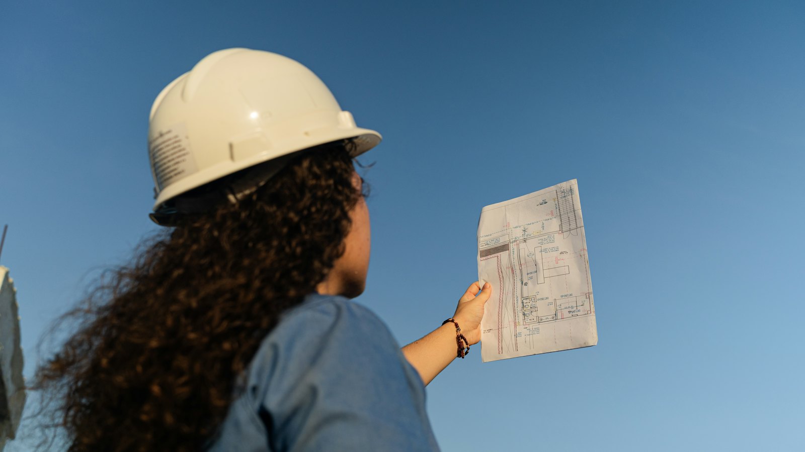 Person in a white hard hat with curly hair, seen from behind, holding a printed plan up against the blue sky