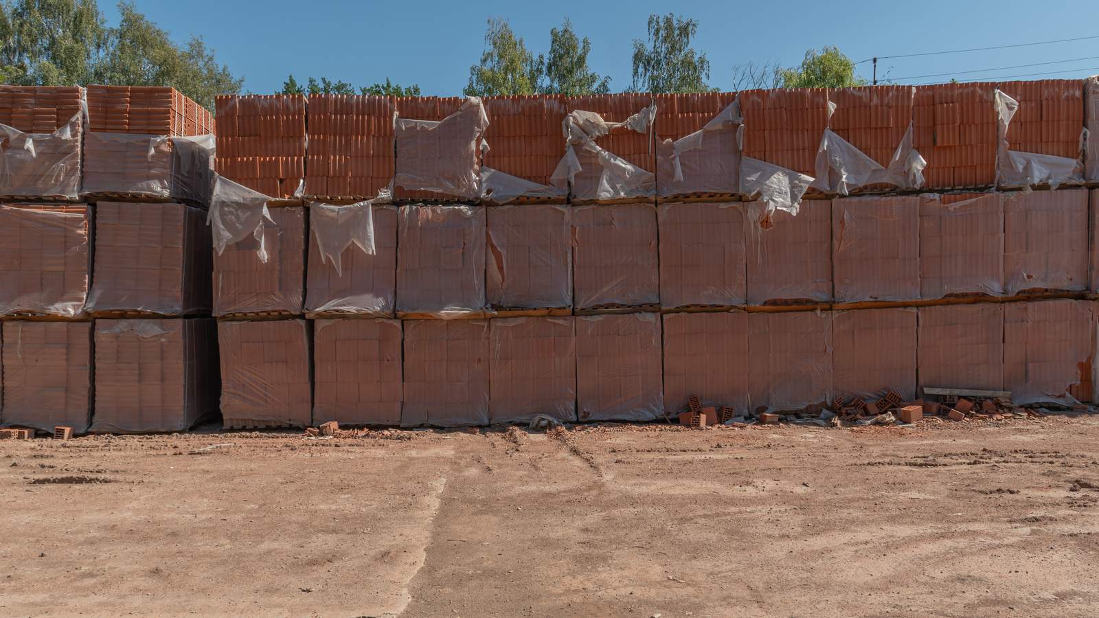 Palés de bloques cerámicos envueltos en film plástico, apilados en dos alturas en un patio de tierra, con bloques rotos junto a la base