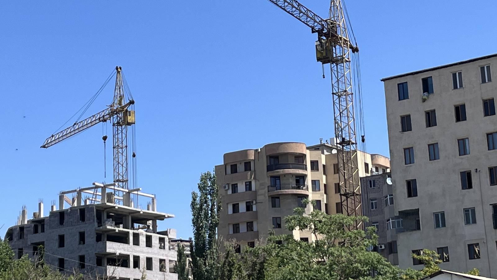 Duas gruas sobre prédios em construção, com a estrutura de concreto de um deles ainda aberta, sob céu azul