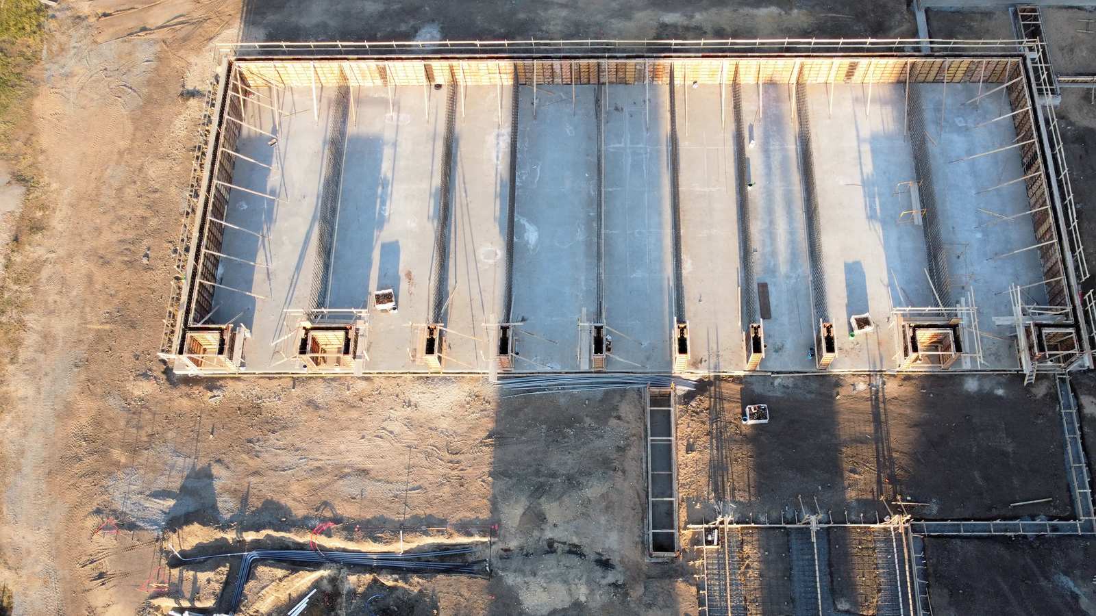 Vista aérea de una cimentación de hormigón en ejecución, con encofrados, armaduras de espera y conductos en la zanja contigua
