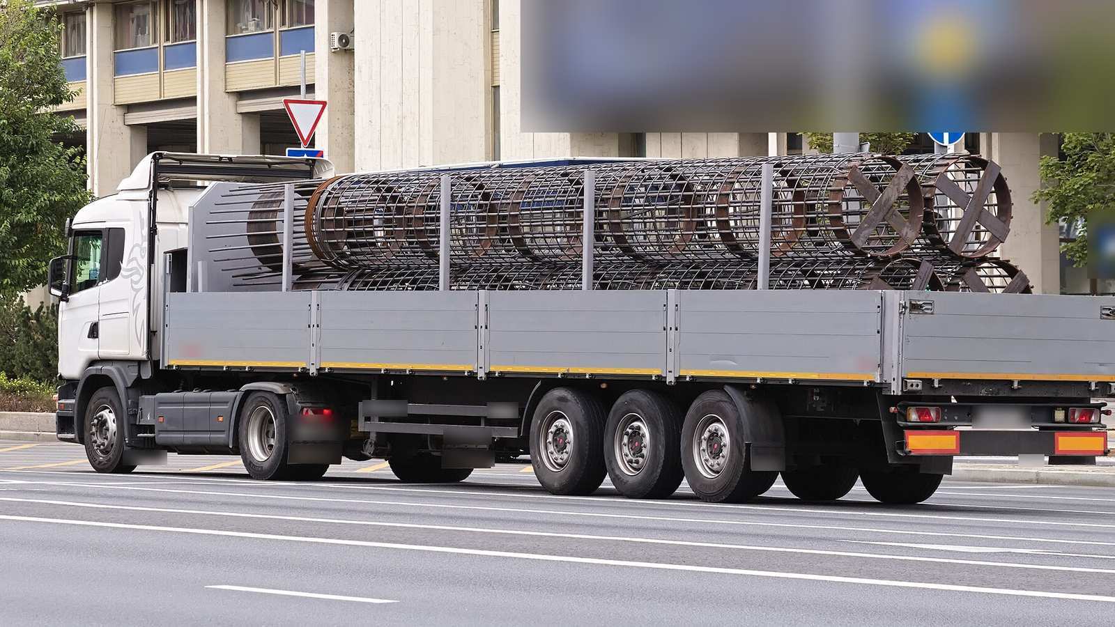 Caminhão com semirreboque de carroceria aberta leva gaiolas cilíndricas de armadura de aço por uma avenida; placas e letreiros borrados